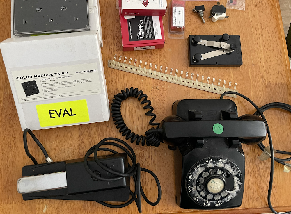Photo of a bunch of items laid out on a table: a rotary telephone, a telegraph device, some LED lights, some AdafruitFeather components, etc